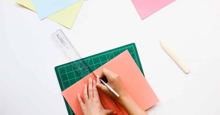 A white surface with a hand holding a ruler and about to draw something on a drawing or cutting mat