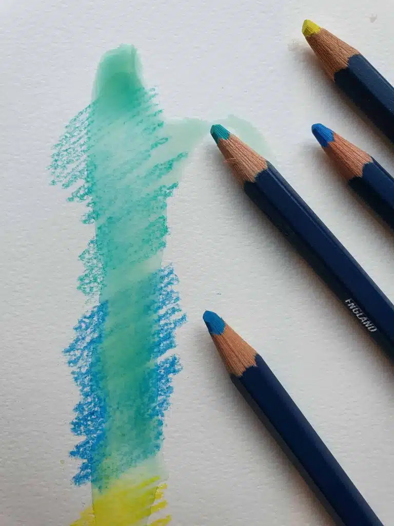 Watercolor pencils blue, green and yellow with a close up of the pencil coloring on the left of them. Water has been applied on top of the watercolor pencils.