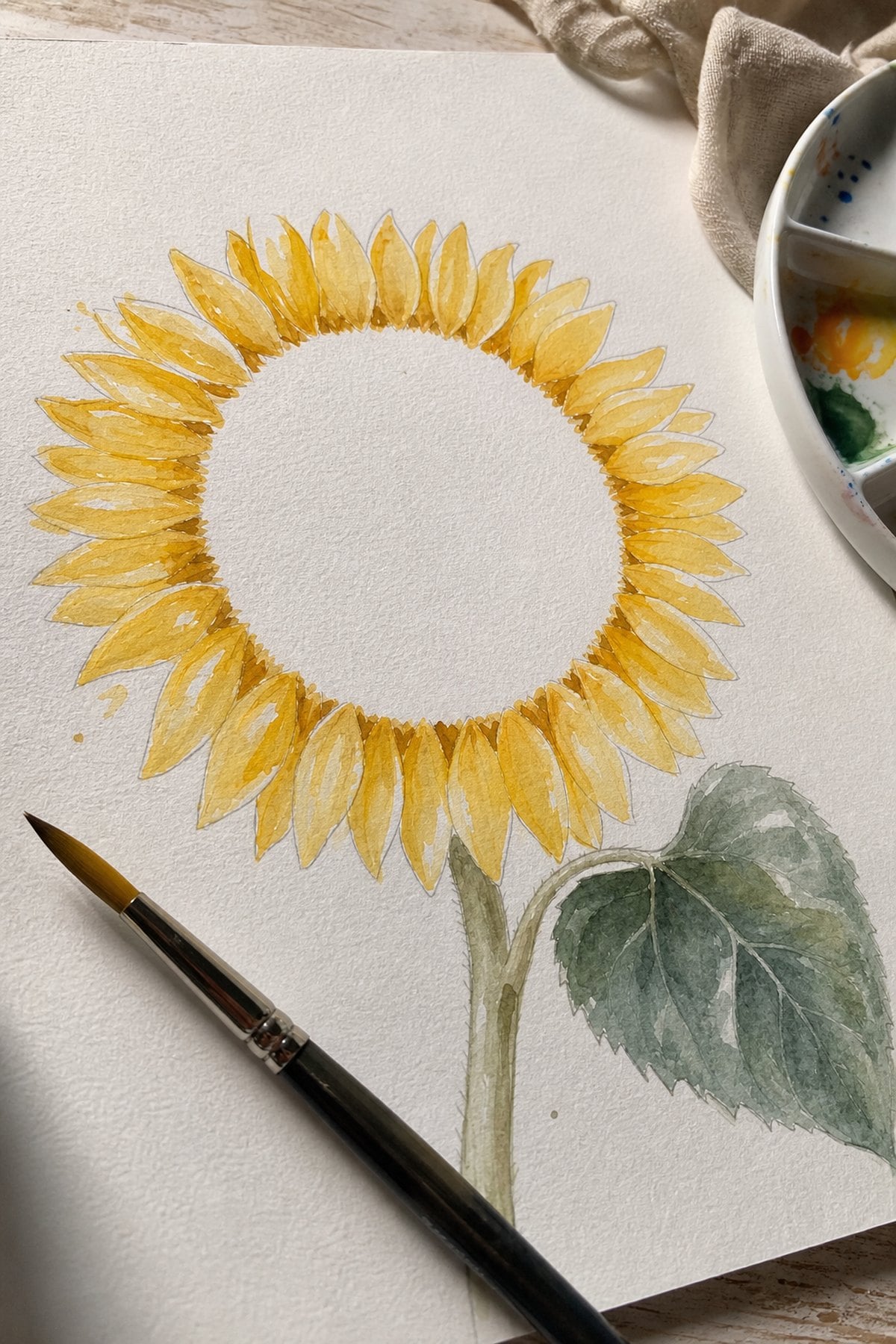 A half-finished watercolor sunflower with the petals washed in golden yellow and the seed centre still bare white paper