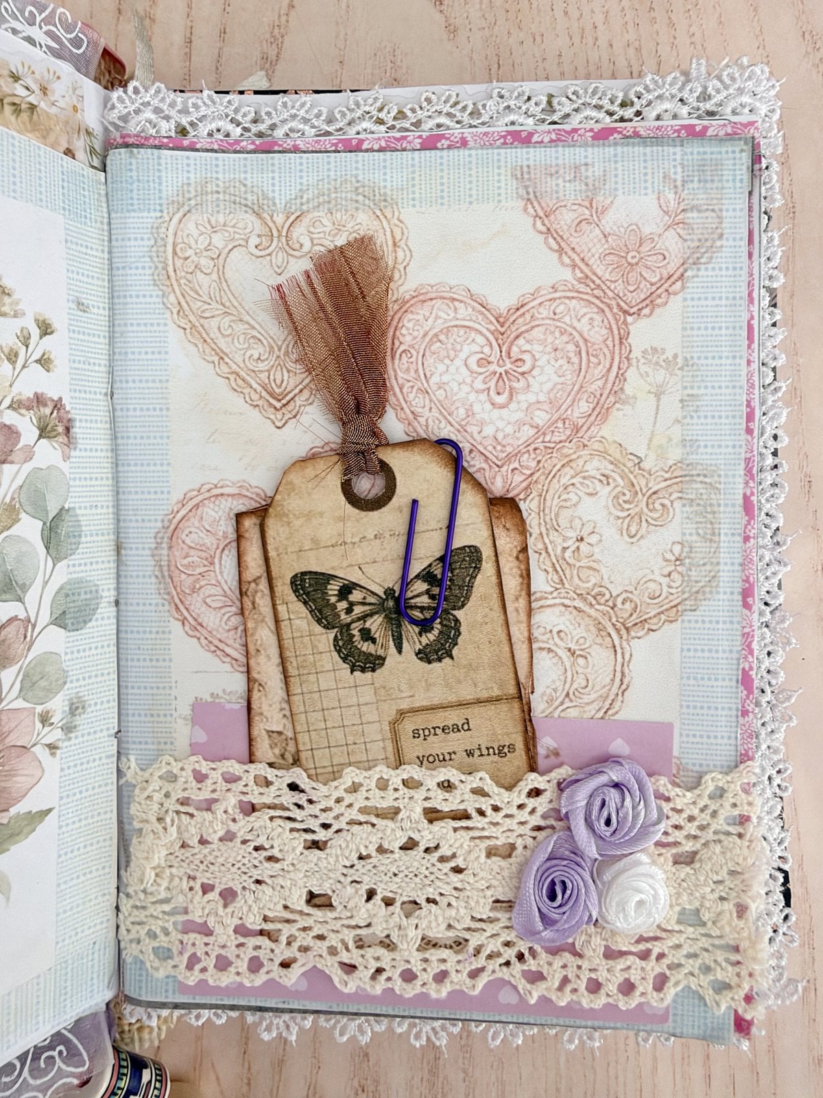 Cream lace pocket with ribbon roses holding a butterfly tag on a pastel page