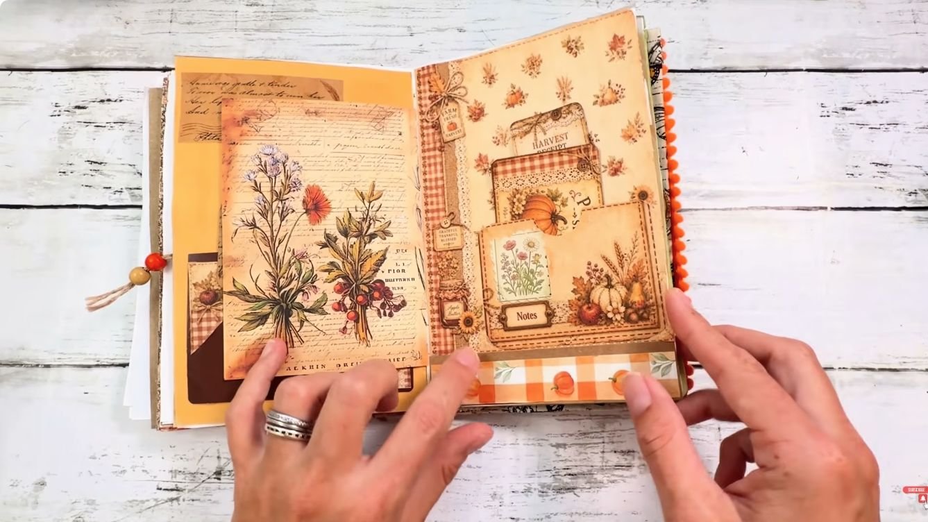 Hands opening a junk journal spread with layered botanical and pumpkin pockets