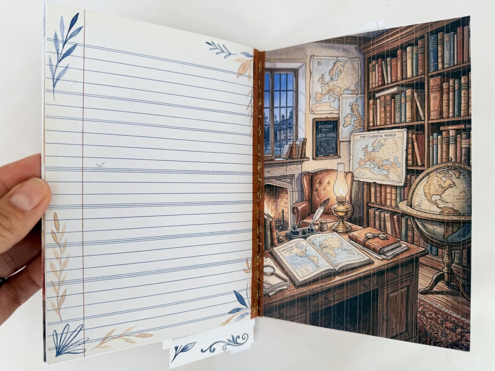 Open mini junk journal with a lined page edged in watercolour leaves on the left and a cosy vintage study scene with books, a globe and maps on the right