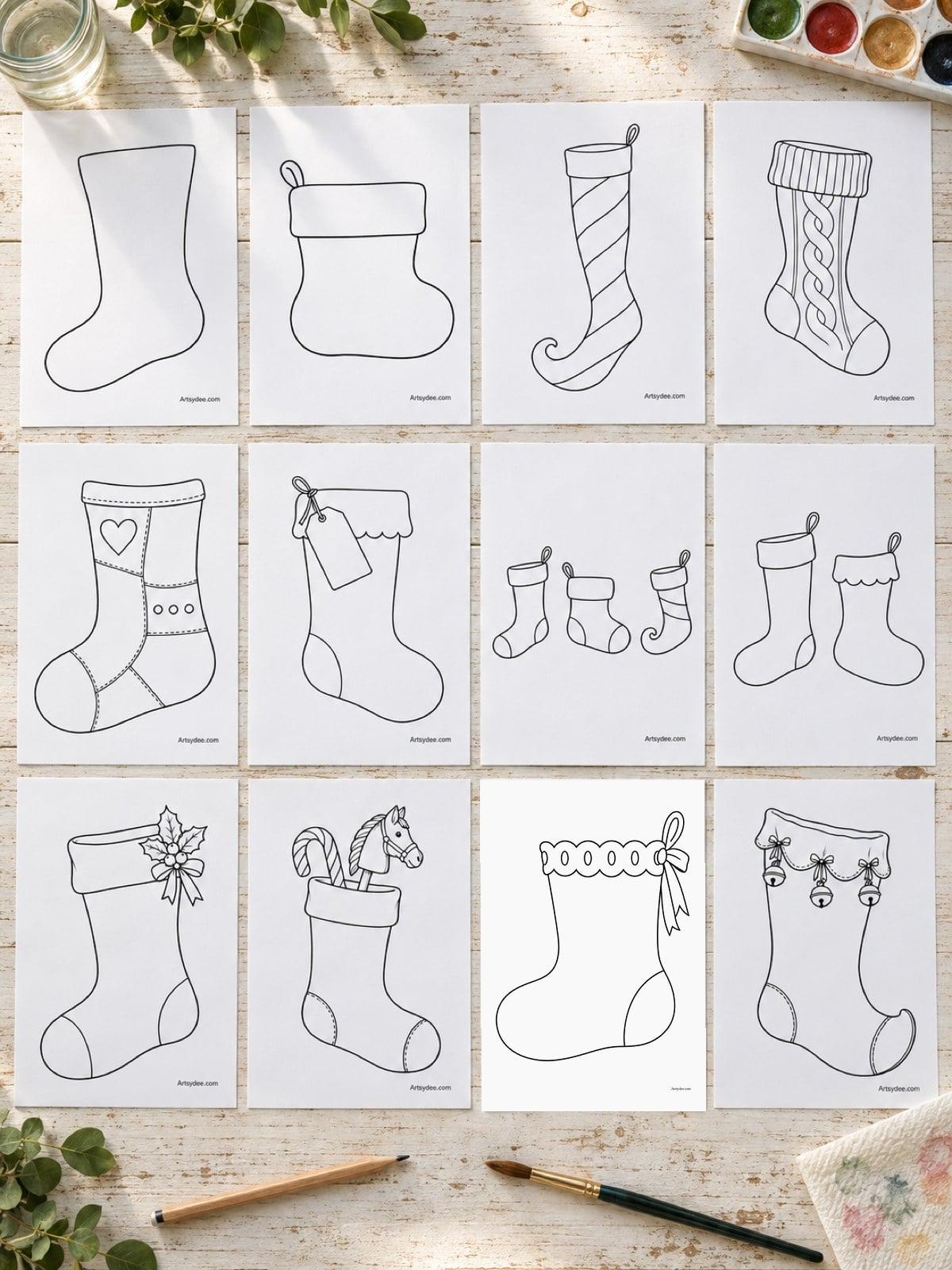 Illustrated mockup of all 12 Christmas stocking templates printed and spread out on a wooden desk with a paintbrush and a watercolour palette