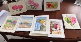Six hand-painted watercolour greeting cards laid out on a table, with pink flowers, strawberries, a lighthouse by the sea, a bird on a branch and a pink heart, by reader Carol S.